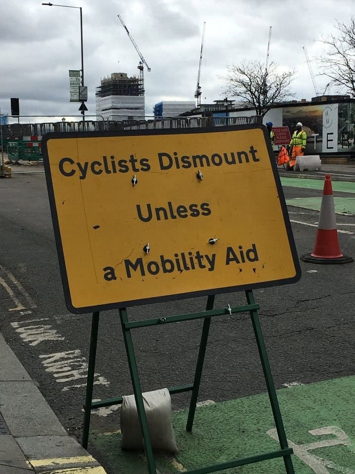 Photo of road works. A large rectangular yellow sign has black text that reads "Cyclists dismount unless a mobility aid"