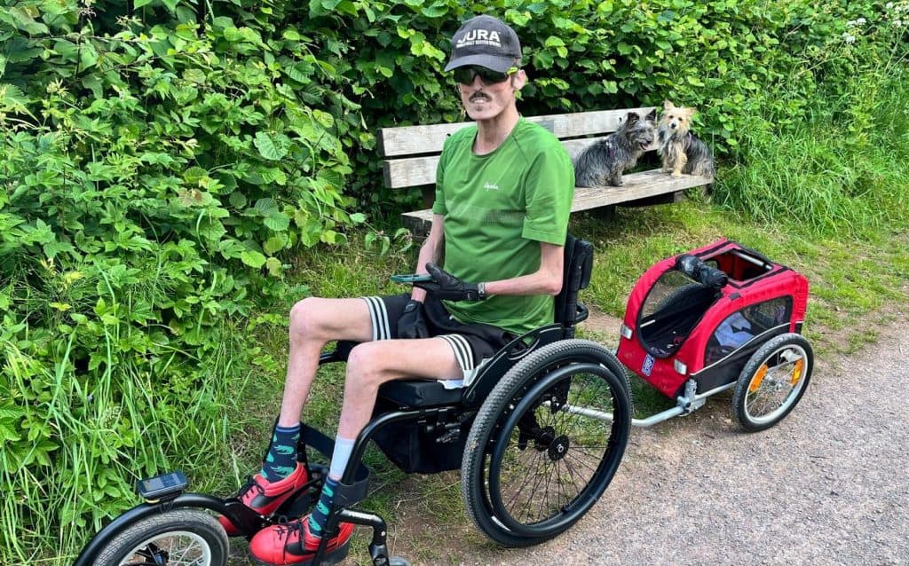Tom in his wheelchair with freeride attachment. He is towing a small trailer. On a bench behind him are two small terriers.