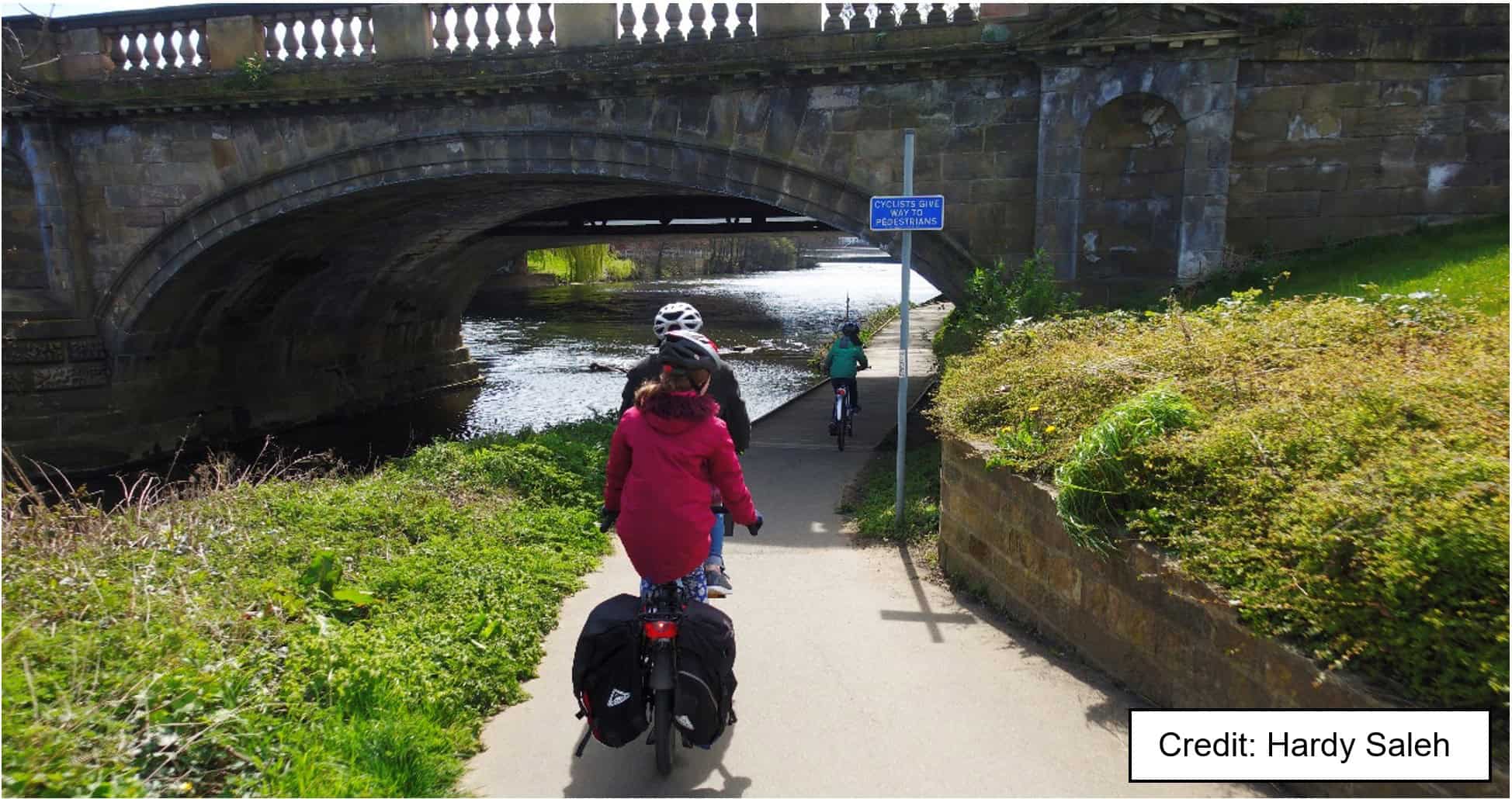 Photo credit Hardy Saleh: A shared cycle and pedestrian path passes under an old stone bridge alongside a river. Under the bridge, the path is a boardwalk about 2m wide with no edge protection. A child is riding a bicycle on the boardwalk, followed by an adult and another child on a tandem. A blue sign reads "cyclists give way to pedestrians".