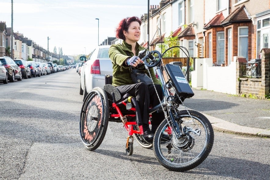 Photo of a handcyclist using a manual wheelchair with clip-on e-handcycle coming towards the camera while turning a gentle corner to their left. The handcyclist's right arm is outstretched even though the crank handle they are holding is most of the way towards their body. Their left hand appears to be holding the downtube - they appear to be gliding around the corner, not pedalling.