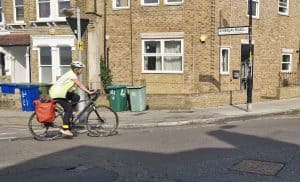 Landscape pic with a side view of Beata cycling on her road bike with orange panniers on a London road. She has dark trousers, a high-viz vest and calf bracelet, pink trainers, and a white helmet 
