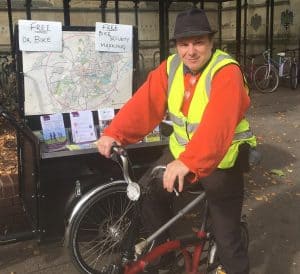 Pic of Ben astride a red Brompton. He is wearing dark trousers, an orange jumper, a hi-viz vest and his customary trilby!