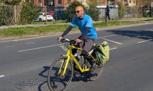 Side pic of Charlie riding his yellow specialized sirrus hybrid bicycle and green panniers on a generic London road. He has grey trainers and cargo shorts and a bright blue t-shirt, with black arm and legwarmers.