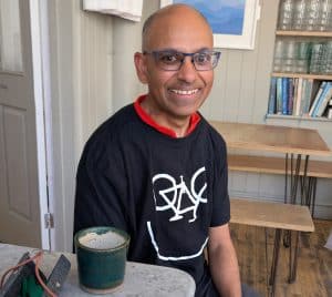 Pic of Charlie in a cafe, with a cappuccino on the white table in front of him. He is smiing, with a mediterranean complexion and stylish glasses. He has a dark blue t-shirt which features an upside down bicycle and a line underneath, so it the graphic looks like a smile.