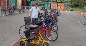 Pic of Charlie on a wide concrete expanse, among shipping containers. He is surrounded by a fleet of non-standard cycles of all shapes and sizes. He has dark trousers, a grey polo shirt, and a black baseball cap with dark sunglasses, and is smiling at the camera