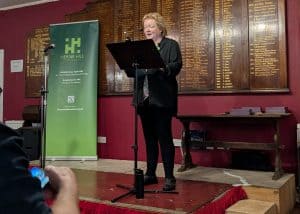 Rebecca Tees standing on the podium addressing the audience. She has medium-length blonde hair and is wearing dark semi-formal clothing. Behind her is alarge green sign for the Herne Hill Society Excellence Awards,