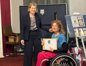 Isabelle receiving our award certificate from Helen Hayes. Isabelle is wearing a turquoise blue loose top and pink trousers and is sitting in her wheelchair. Helen Hayes MP, in a dark trousersuit and green/grey patterned blouse, is to her right