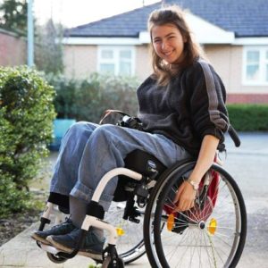 Pic of Katie smiling as she performs a wheelie in her silver wheelchair. She is wearing loose denim trousers, grey socks and deck shoes, and a black jumper. She has long brown loose hair.
