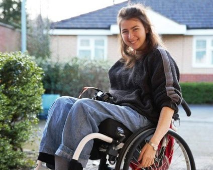 Pic of Katie smiling as she performs a wheelie in her silver wheelchair. She is wearing loose denim trousers, grey socks and deck shoes, and a black jumper. She has long brown loose hair.