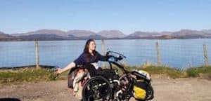 Pic of Katie on the banks of a beautiful loch. She is in her wheelchair with clip-on hybrid handcycle attachment, which is fully laden for touring!