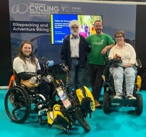 Katie at the National Cycling Show after delivering a panel discussion. She is in her wheelchair with clip-in hybrid hand-cycle attachment and attached luggage. With her are two gentlemen standing and a lady in a powerchair, All are smiling