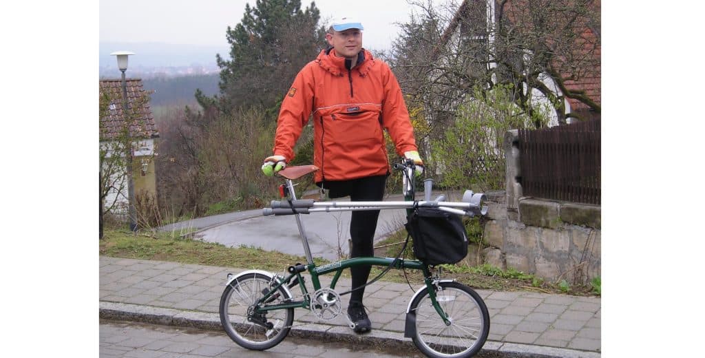 Pic of Kevn standing on the pavement at the side of a road. He is behind a green Brompton folding bicycle, to which he has attached two crutches. He has one leg, wearing winter cycling tights and an orange waterproof smock. He has yellow winter cycling gloves and a blue and white cycling cap.