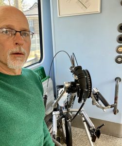 Pic of Kevin on the train. Kevin has glasses and a white short-trimmed beard. He is wearing a green jumper. Behind him and slightly to the right is a Stricker handcycle attachment