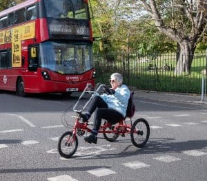 A person is cycling over an on-road cycle crossing on a red semi-recumbent trike, in front of a red double-decker bus. There is a park with black iron railings behind them and the bus.