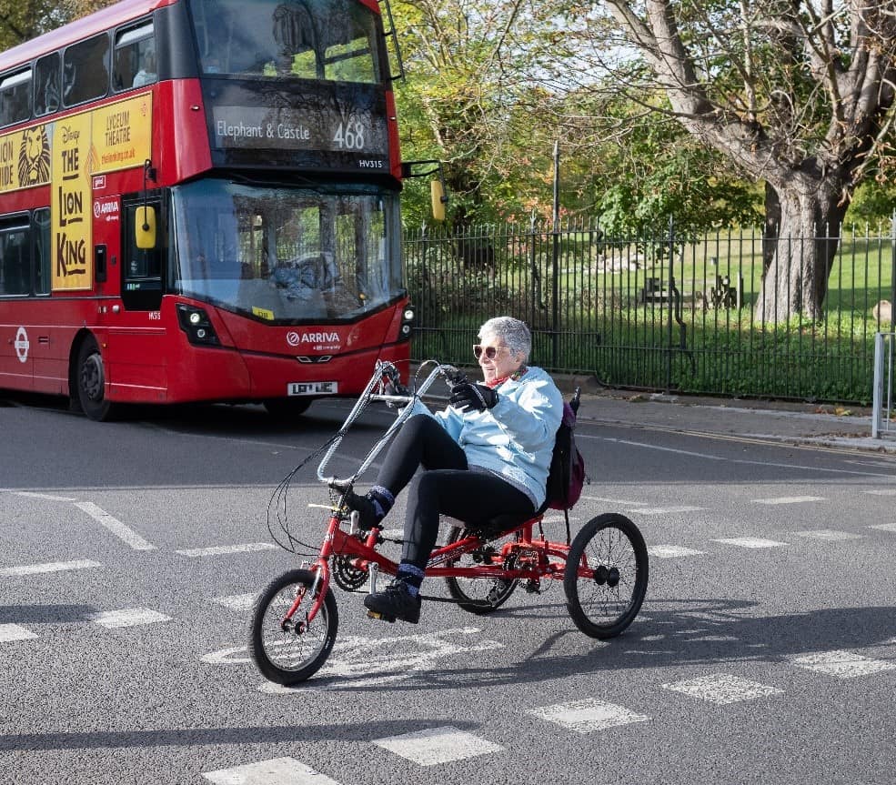 A person is cycling over an on-road cycle crossing on a red semi-recumbent trike, in front of a red double-decker bus. There is a park with black iron railings behind them and the bus.
