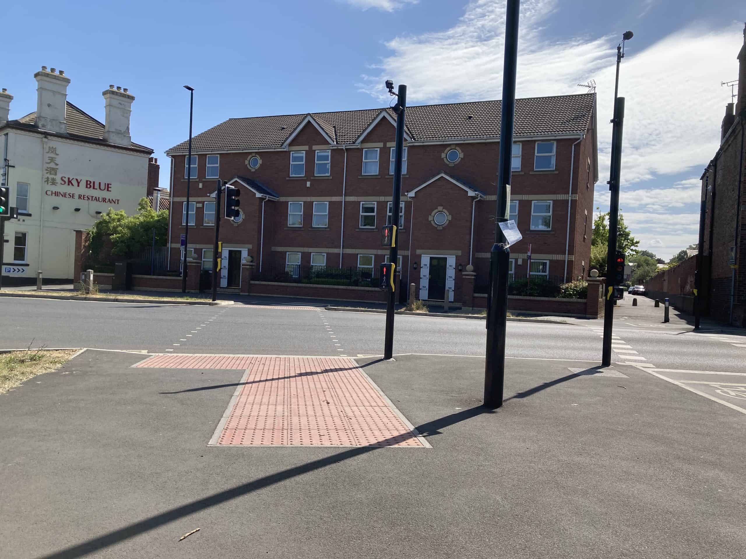 A wide pedestrian crossing going straight across a road but then requiring pedestrians to stop on a refuge to cross a cycle track. A cycle track crosses the carriageway to the right of the pedestrian crossing and separately from it.