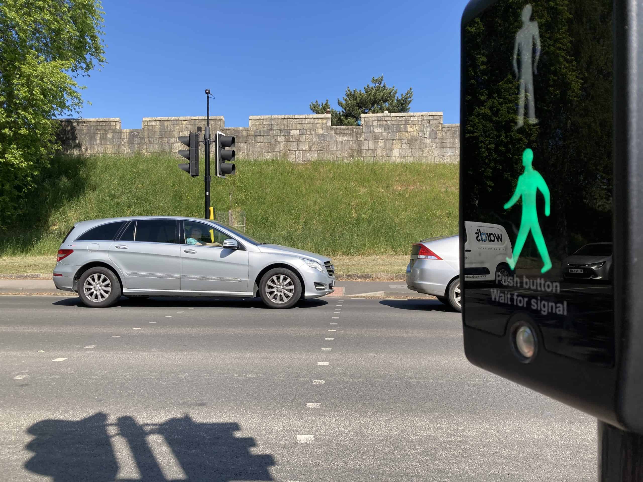 A pedestrian crossing over a busy road. The green man is lit showing pedestrian right to cross, but a car has stopped blocking the dropped kerb on the far side of the road.