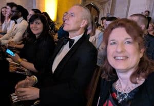 Picture of Isabelle in the audience at an awards evening, She is wearing a midnight blue and red patterned dress and necklace, and has worn her red/brown hair long. She is smiling at the camera. In the background is a gentleman in a tuxedo with a shaved head, and a younger lady in a black dress. They are all looking up at the stage (out of shot)