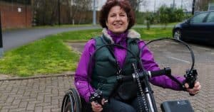 Upper-body image of Isabelle using her wheelchair with clip-on handcycle attachment. She is wearing a purplelong sleeve top and puffa gilet, and looking at the camera. In the background, a small grassy area and road can be seen in a residential area,