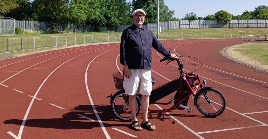 Image of Eric standing next to a red cycle with one hand holding the handlebar. He is on a red sports track with a metal fence, grass and trees in the background. He is smiling at the camera and is wearing glasses, a blue shirt, white shorts, brown sandals and a white hat. 