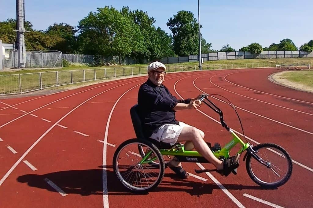 Image of Eric sat on a green cycle. He is holding the handlebars and looking at the camera. He is on a red sports track with a metal fence, grass and trees in the background. He is wearing glasses, a blue shirt, white shorts, brown sandals and a white hat. 