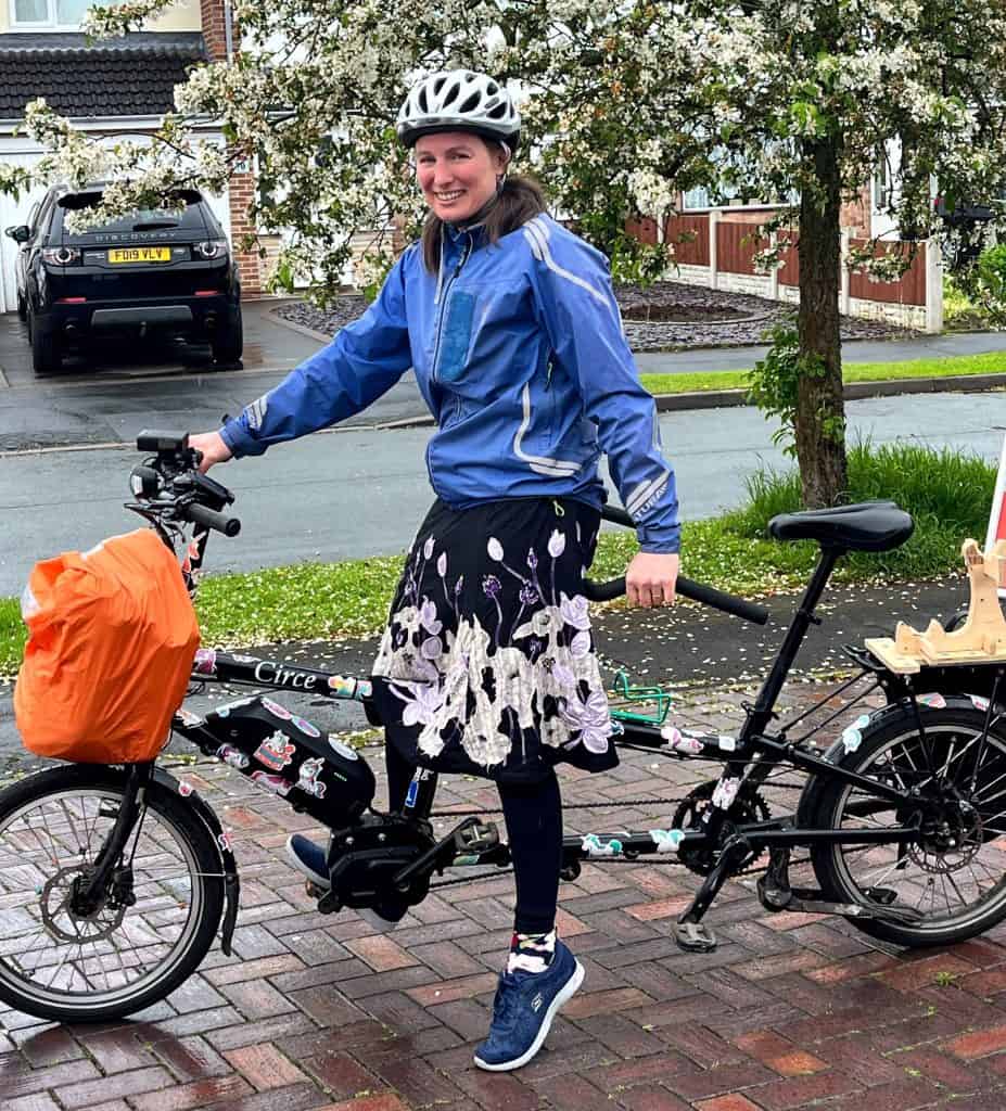 Kate Bell sat on a tandem cycle. She is a white woman with brown hair and is wearing a white cycling helmet, a blue jacket, dark blue skirt with white flowers and blue trainers. The bike has an orange bag at the front. In the background is a tree with white flowers, a road, and a car opposite parked in a driveway.