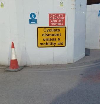 Photo of white painted hoardings around a work site on road. There is a red "Cyclists Dismount And Use Footway" sign. Below this, there is a yellow "Cyclists dismount unless a mobility aid" sign.