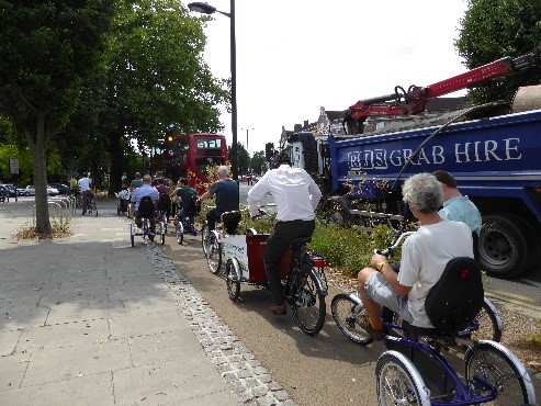 Photo of lots of people riding non-standard cycles largely in single file on (and off) a very narrow cycle track that is edged with setts then a wide footway to the left and green planting then a busy road with a grabber truck and double decker bus to the right.