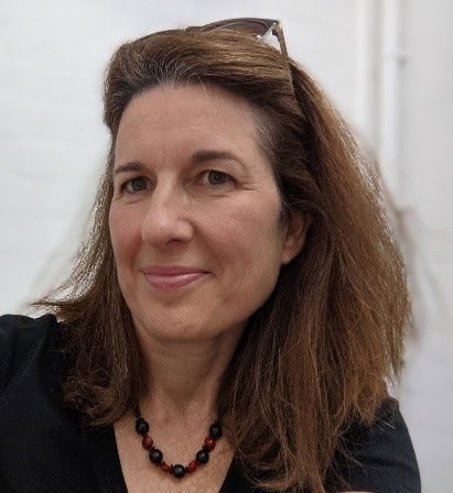 Head and shoulders photograph of Isabelle Clement: a white woman with long, straight auburn hair. She is wearing dark open neck shirt and a dark bead necklace. She has sunglasses pushed onto the top of her head and is smiling and looking slightly away from the camera. 