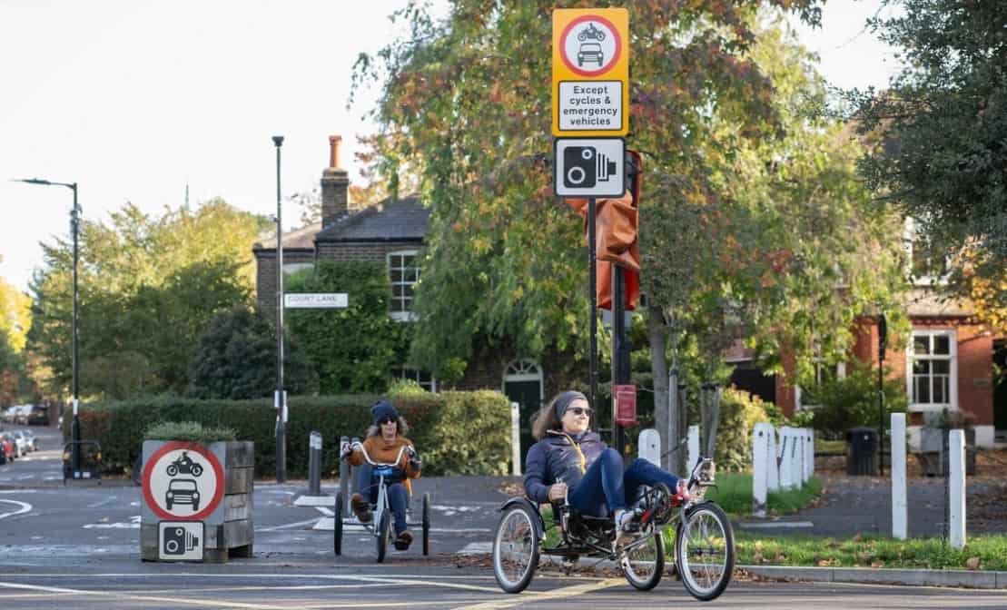 Photo of a CCTV-enforced modal filter on a sunny and leafy residential street. Signs say no vehicles except cycles and emergency vehicles. Two people are riding through the modal filter on tricycles.