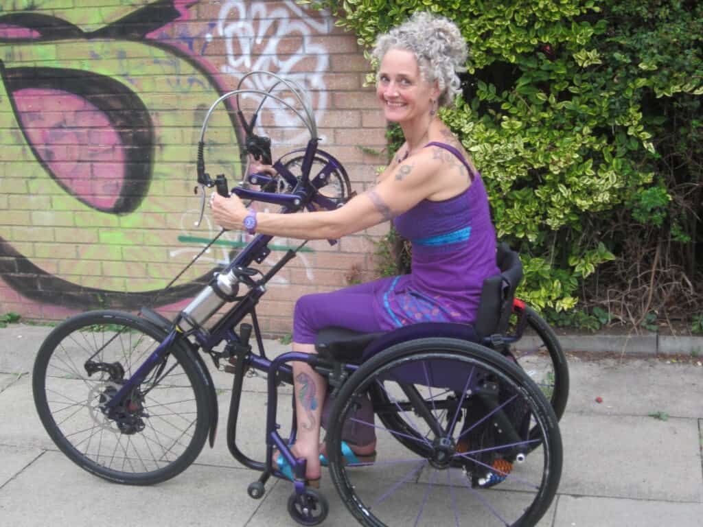Photo of Kay on her DaVinci e-assist clip-on handcycle. She is wearing a purple top and is smiling at the camera. In the background is a wall covered with graffiti.