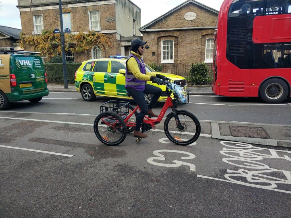 A black woman on a red upright tricycle cycling in a cycle lane. The cycle has a basket in which is a blue bag. She is wearing a black helmet, yellow long-sleeved top and a purple high vis jacket. Behind her is a red bus, yellow and green ambulance and a green van. In the far background are houses.