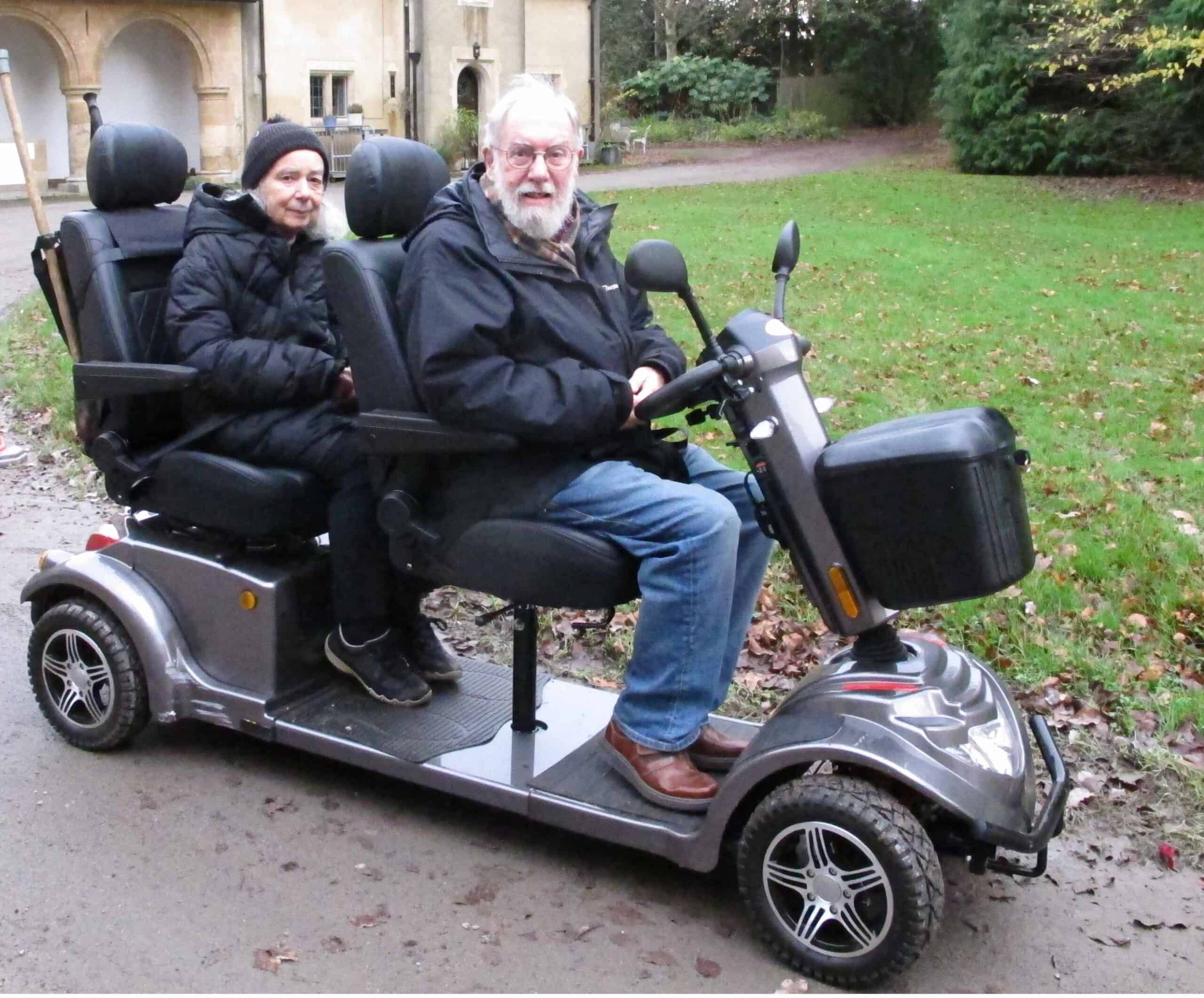 Two people sitting one behind the other looking at the camera. They are on comfy-looking black seats on a silvery grey tandem mobility scooter in a grassy churchyard