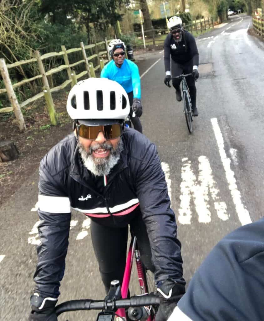 A photo of Mark Browne. He is a black man with a beard riding a bike on a road with white road markings. He is wearing a white helmet, sun glasses and a blue long-sleeved jacket. He is smiling at the camera. Behind him are two other cyclists - a black woman wearing a bright blue top and white helmet and a black man wearing a white helmet and black top. To the left is wooden fencing 