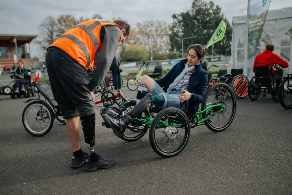 A picture of an inclusive cycling session. There is a man with an artificial leg wearing a high vis jacket learning over looking at a recumbent tricycle. There is a white woman in it with dark hair wearing a blue top and jeans. In the background are more cycles.