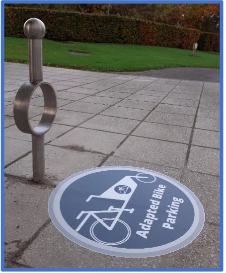 A photo of an accessible cycle park. There is a blue sign in front of it which has a tandem cycle and the words adapted bike parking
