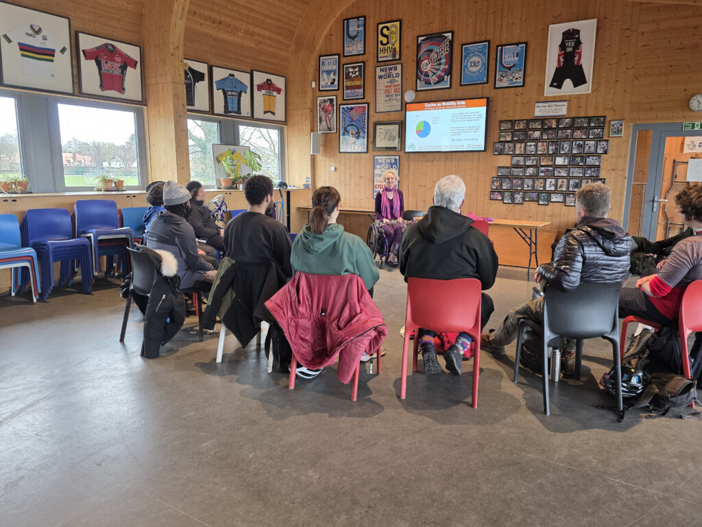 A photograph of a training session inside a room which has wood walls covered photos and framed jerseys. At the front facing the camera is a white woman with blond hair in a wheelchair wearing a pink scarf and a deep purple top and trousers. Behind her head is a screen showing a slide with the title “Cycles as Mobility Aids”. In front of her is a semi-cycle of people watching attentively.
