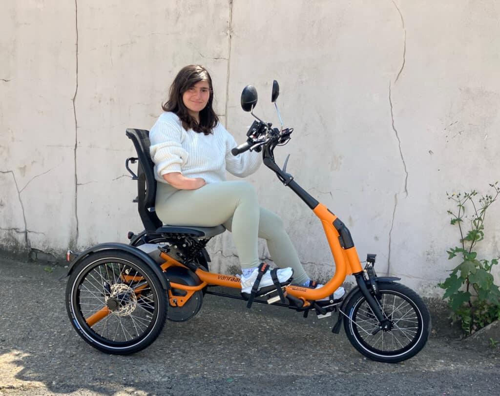 A photo of a white woman with brown hair on an orange vanRaam Compact Easy Rider tricycle. She is wearing a white top and beige trousers and is smiling at the camera. Behind her is a white wall with vertical crack. There is a large green weed growing from the base of the wall just in front of her tricycle.
