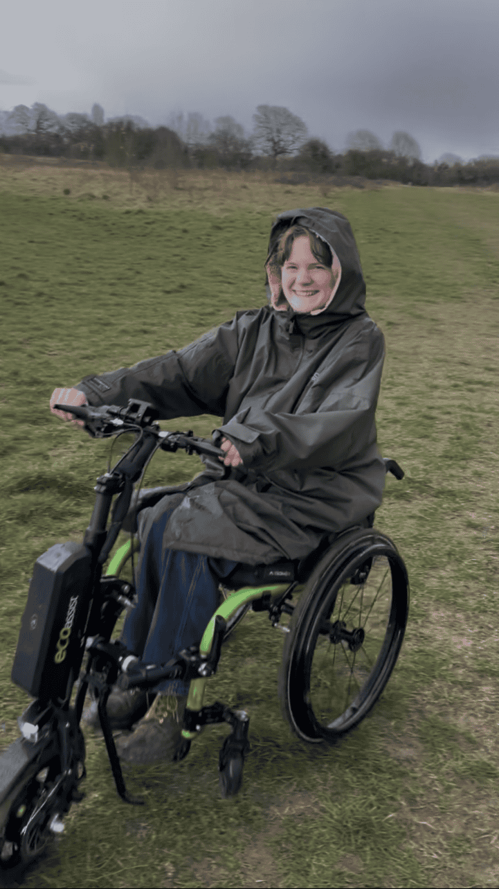 A young white woman smiling at the camera, using a bright green manual wheelchair with front power assist on a grassy open space with trees in the background. She is wearing a big khaki green coat that looks very wet.